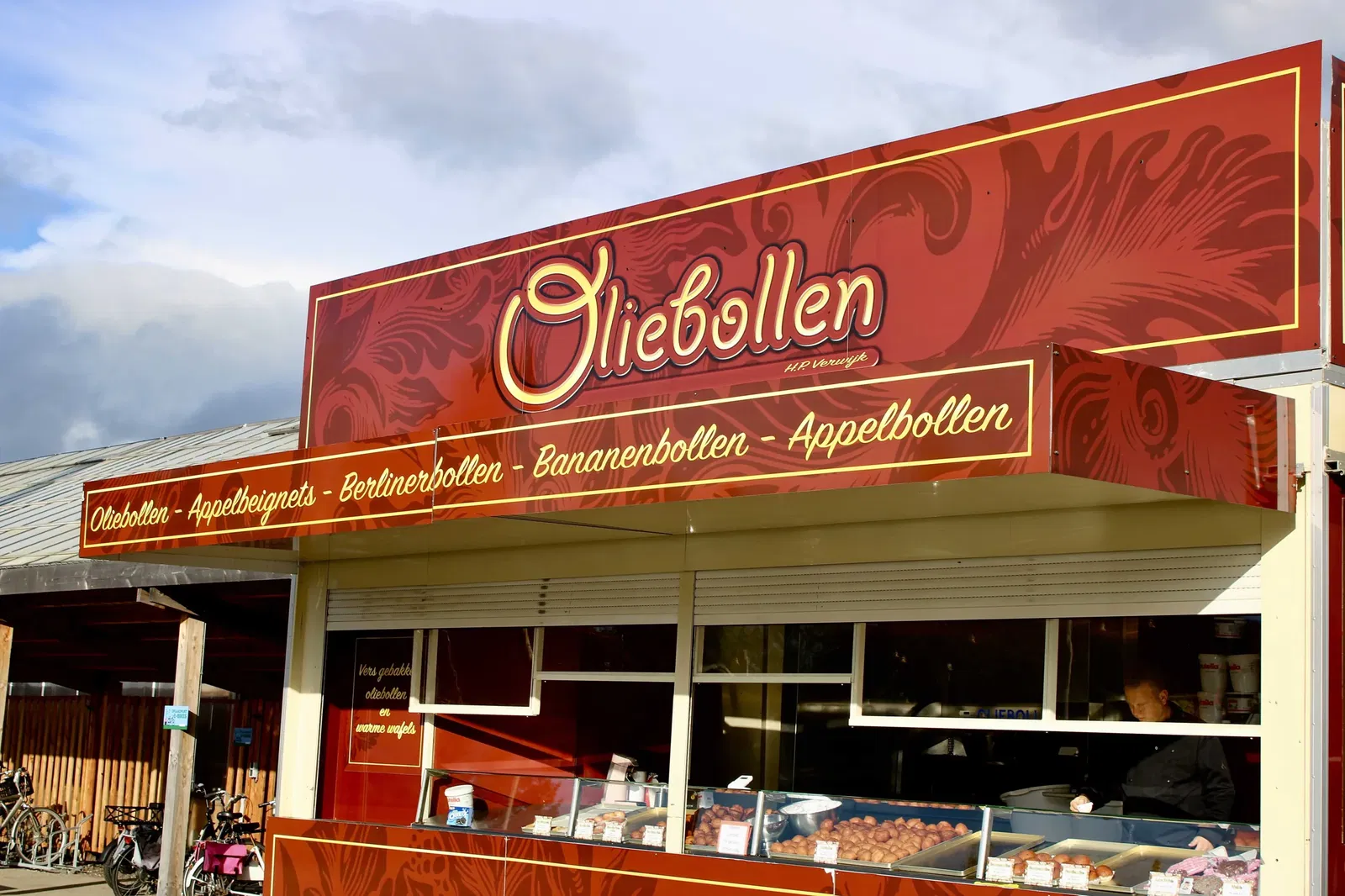 De oliebollenkraam bij Steck van voren gefotografeerd. Boven de bakken met oliebollen, appelflappen en andere baksels zijn rode borden zichtbaar met daarop de woorden 'oliebollen, appelbeignets, berlinerbollen, bananenbollen, appelbollen'