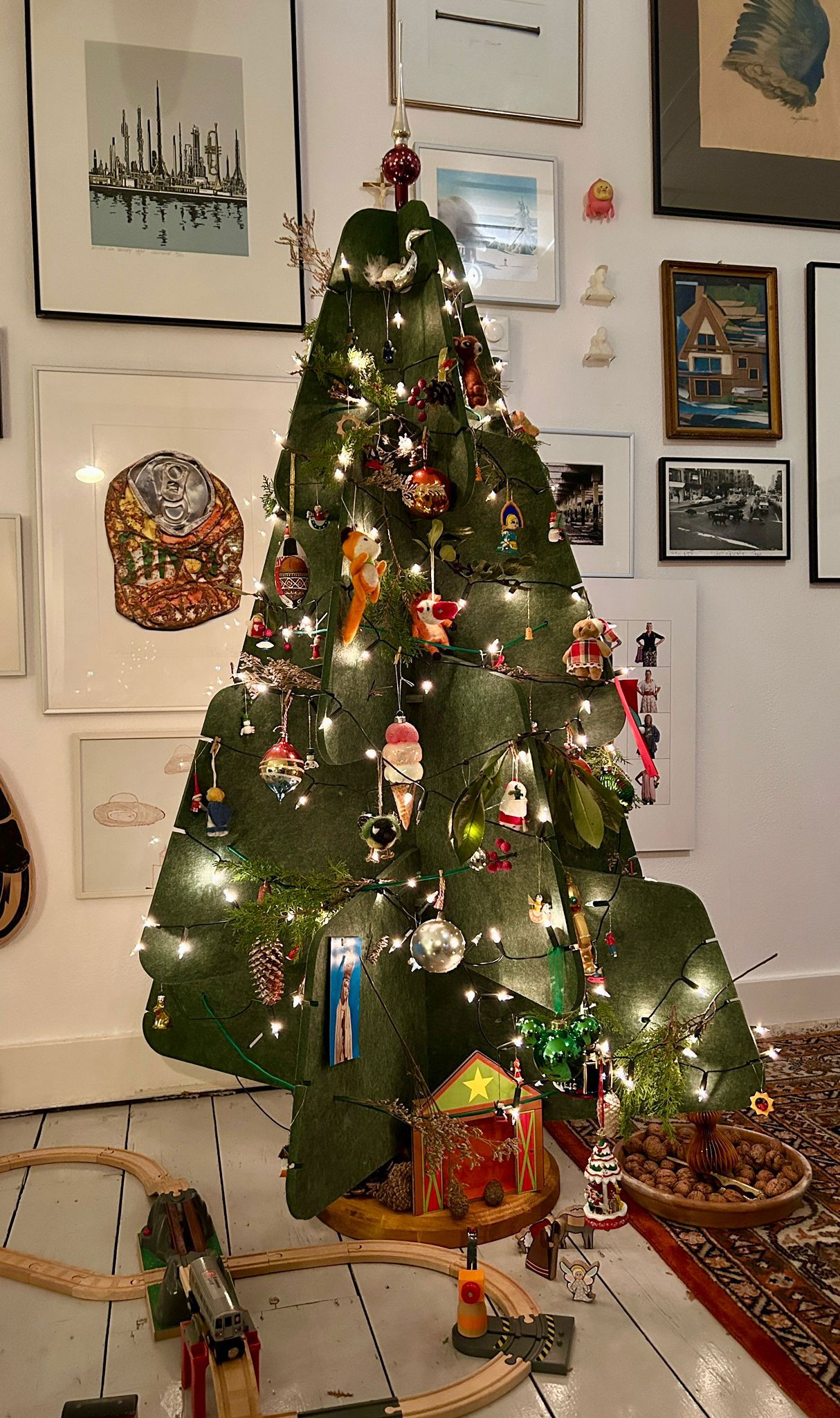 Een groene vilten kerstboom van Jingle Tree staat versierd in een gezellige woonkamer. De boom is gedecoreerd met traditionele kerstballen, kleine figuurtjes en lichtjes die hem een warme sfeer geven. Onder de boom ligt een houten speelgoedtrein en enkele natuurlijke decoraties zoals walnoten.